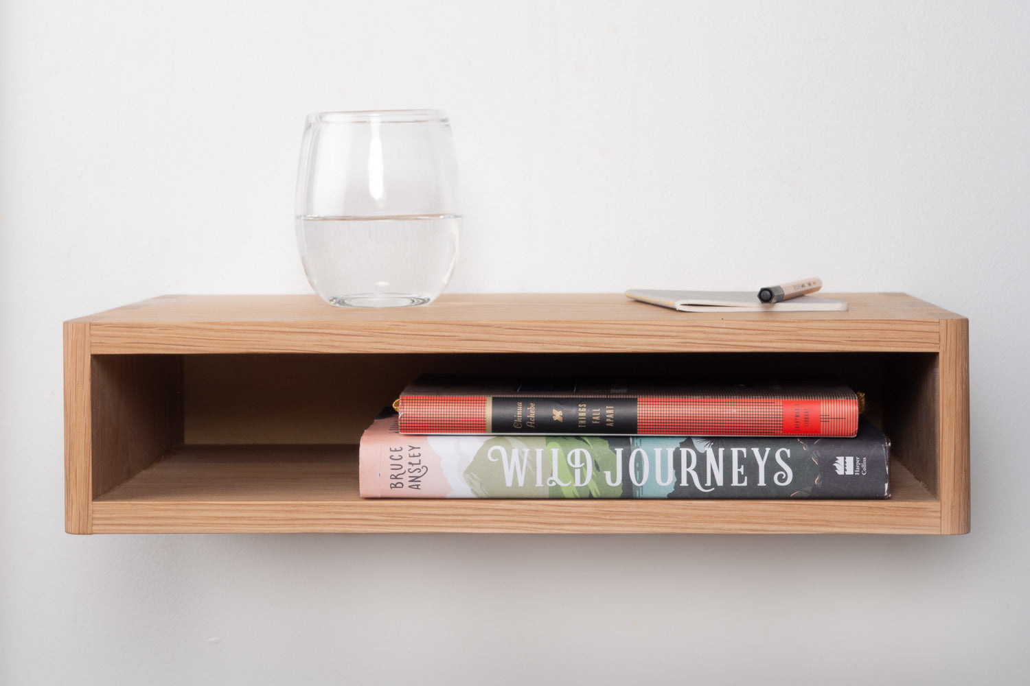 Mesa Bedside Shelf in White Oak pictured with a glass of water, notebook, pen and two books. Designed and made by Hey, Porter.