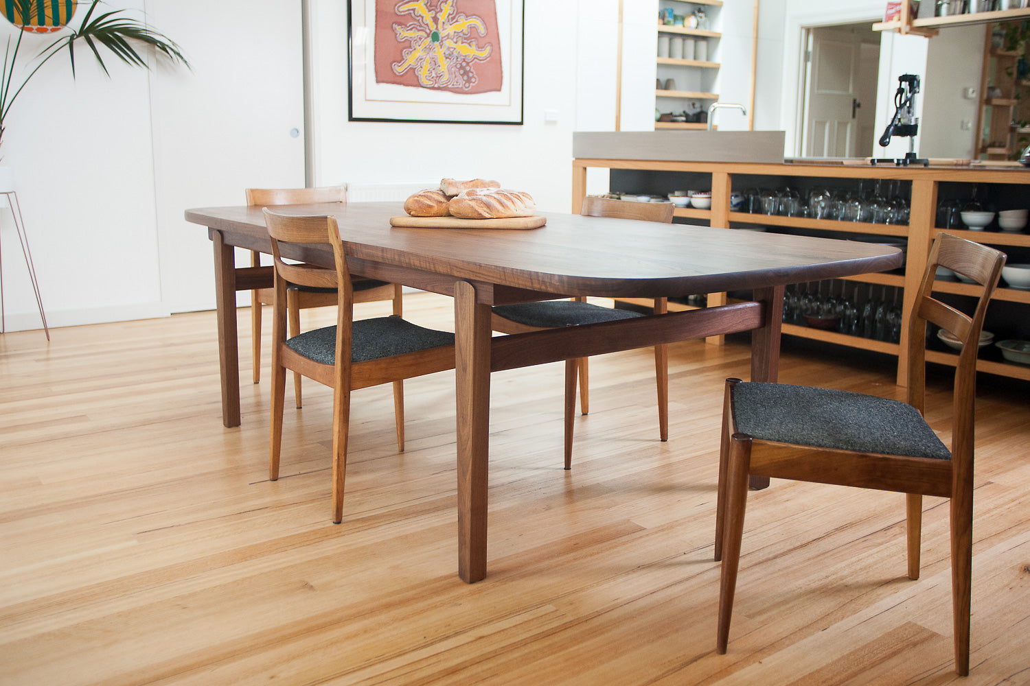 Wesley Dining Table in Walnut pictured with bread, dining chairs with kitchen in the background - 3/4 view. Designed and made by Hey, Porter.