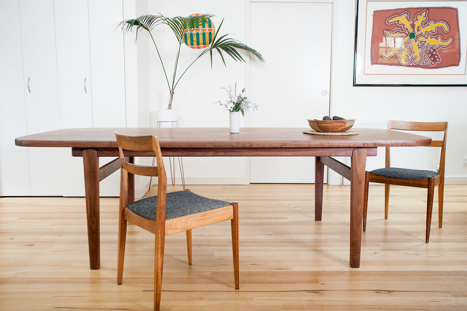 Wesley Dining Table in Walnut with dining chairs, bowl of avocados and plant in background - front view. Designed and made by Hey, Porter.